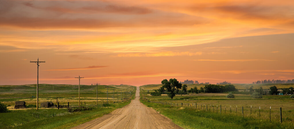 A rural dirt road stretches into the distance under a vibrant orange and yellow sunset sky in Sterling, Colorado. Power lines run parallel to the road, with green fields and scattered trees on either side, creating a peaceful and picturesque countryside scene.