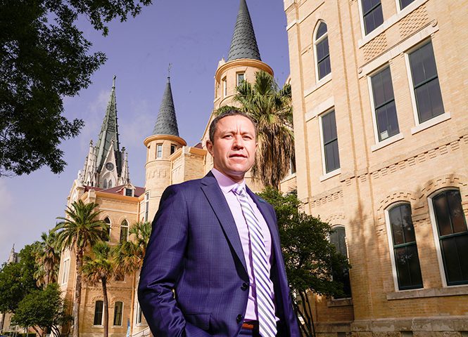 A man in a blue suit and striped tie stands confidently in front of a historic building with pointed towers and palm trees. The building's architecture features a mix of gothic and classical elements. The sky is clear, and the man looks directly at the camera, exuding a sense of professionalism and authority.