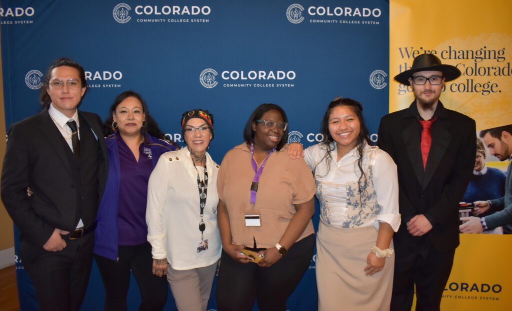 Six individuals stand together in front of a Colorado Community College System backdrop, smiling at the camera. A yellow banner on the right reads, "We're changing the way Colorado goes to college."