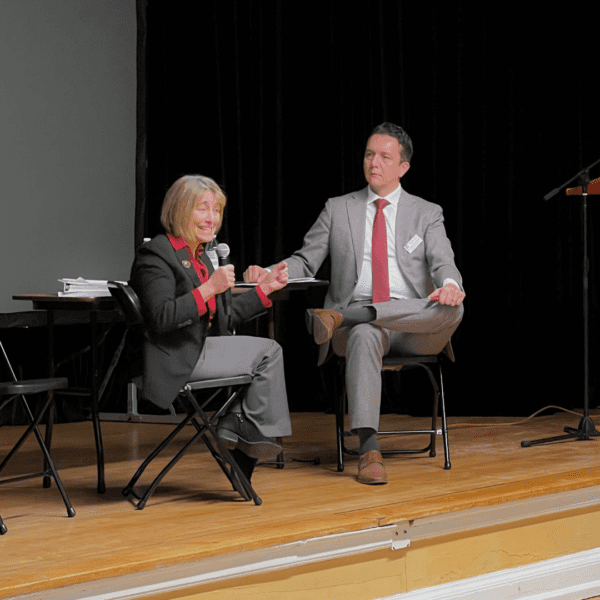 Two individuals sit on stage in conversation; one is speaking while the other listens.