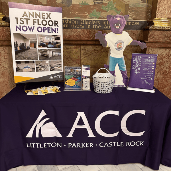 An informational table promoting Arapahoe Community College (ACC) with a purple tablecloth, brochures, a mascot cutout, and signage announcing the opening of the Annex 1st floor.