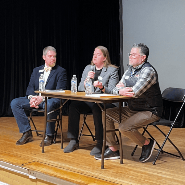 Three panelists sit at a table on stage, one speaking into a microphone while the others listen attentively.