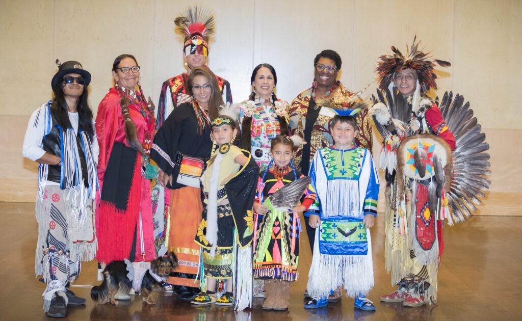 A group photo featuring individuals of various ages dressed in vibrant traditional Indigenous regalia, showcasing intricate patterns, beadwork, and feathers. They are standing together and smiling, with a small dog visible at the front wearing a decorative outfit. The setting appears to be indoors with a plain wall background.