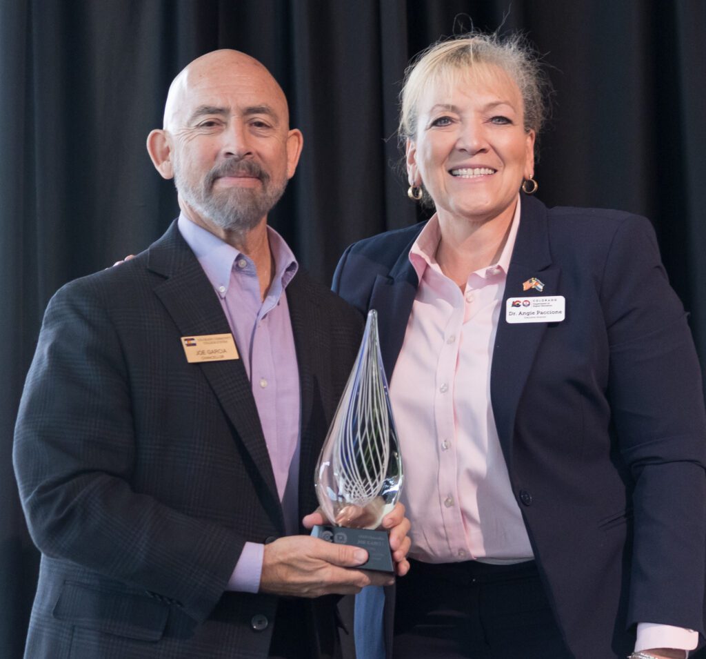 Two individuals standing side by side, smiling. The man on the left is holding a glass award with intricate designs, and he is wearing a suit with a name badge that reads 'Joe Garcia.' The woman on the right, also in a suit with a name badge that reads 'Dr. Angie Paccione,' has her arm around him. They are standing against a dark curtain backdrop.