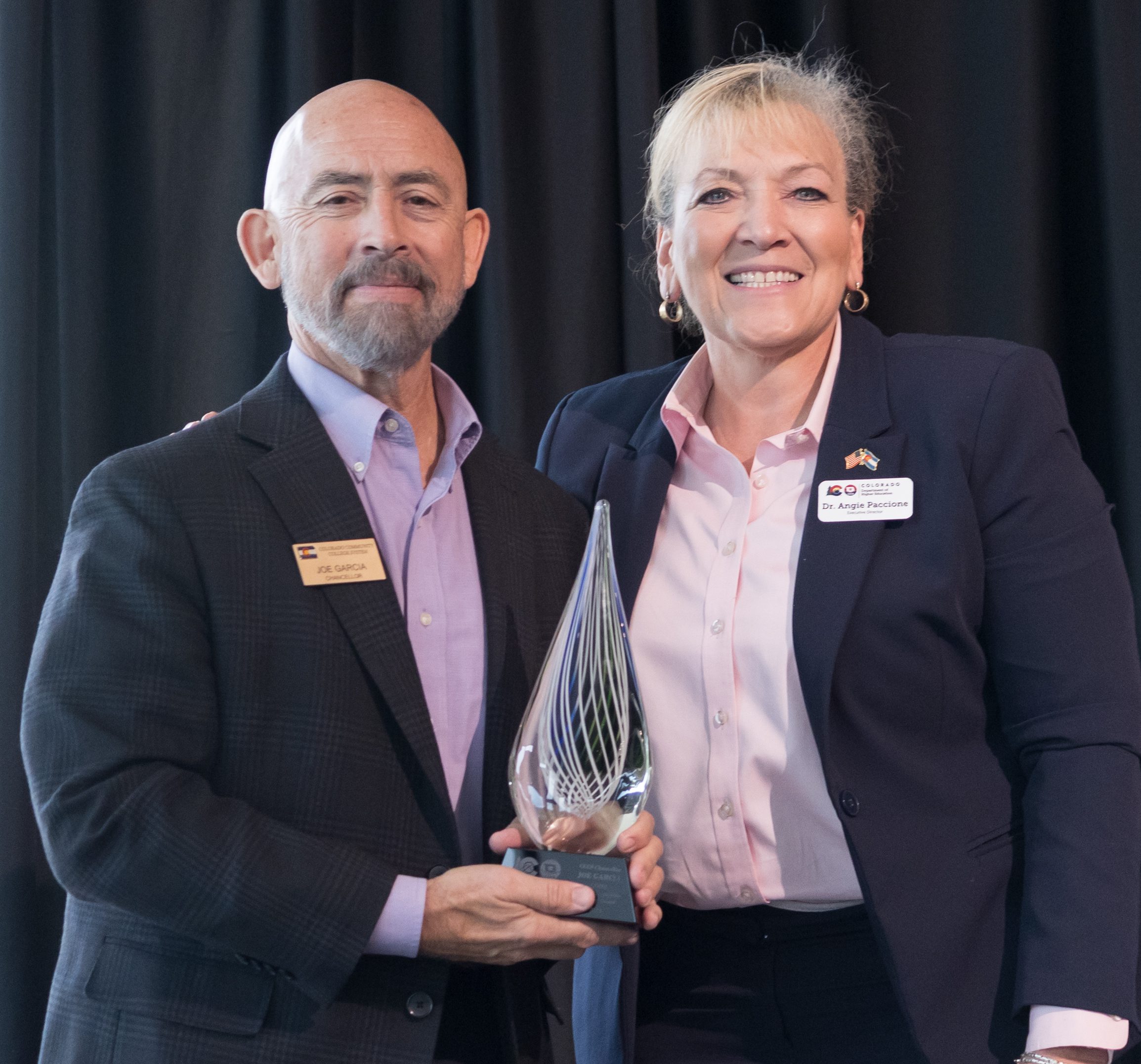 Two individuals standing side by side, smiling. The man on the left is holding a glass award with intricate designs, and he is wearing a suit with a name badge that reads 'Joe Garcia.' The woman on the right, also in a suit with a name badge that reads 'Dr. Angie Paccione,' has her arm around him. They are standing against a dark curtain backdrop.