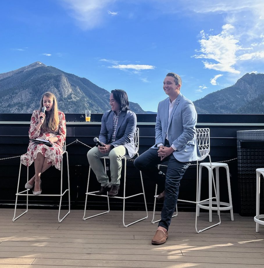 Three people are sitting on tall chairs on an outdoor deck, participating in a panel discussion. A woman on the left, holding a microphone, is speaking. The other two panelists, a man in the center and another man on the right, are listening and smiling. Behind them, there is a stunning mountain backdrop with clear blue skies.