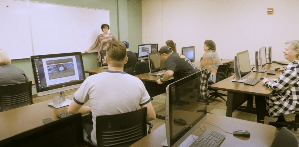 A classroom setting with students seated at desks, each with a desktop computer. A teacher stands at the front near a whiteboard, addressing the class. The room has a professional yet casual atmosphere, with students of various ages engaged in learning. The visible computer screens display educational content related to cybersecurity.