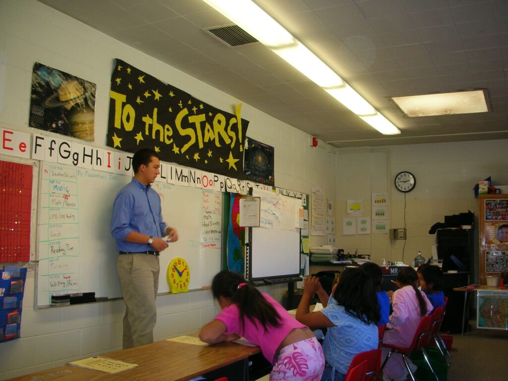 A teacher stands at the front of a colorful classroom, engaging students seated at desks. A “To the Stars!” banner hangs on the wall.